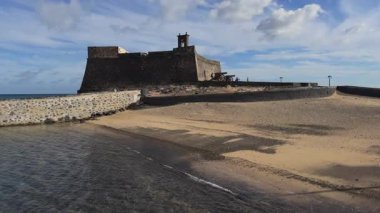 Taş kaldırım tarafından ulaşılmış nefes kesici bir deniz kenarı şatosu, tarihi savunma rolünü vurgulayarak yavaş çekim dalgalarıyla sergileniyor..