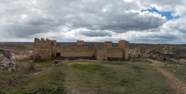 Çarpıcı gökyüzünün altında, kırlara ve uzak bir kasabaya bakan heybetli antik taş bir kale var..