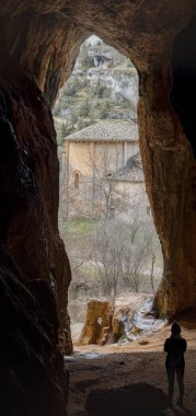 Birisi bir mağarada duruyor, kayalık bir arazi ve çıplak ağaçların ortasındaki antik bir şapele bakıyor..
