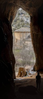 Mağara girişindeki bir insanın silueti engebeli uçurumların ve seyrek ağaçların arasındaki antik bir taş binayı görüyor..