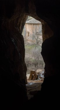 Tarihi bir kilise, çıplak ağaçlar ve kaya oluşumlarıyla çevrili huzurlu bir manzarayla çevrili bir mağara girişinden görülür..