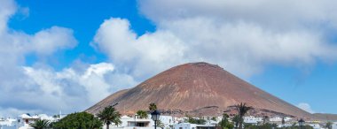 Lanzarote 'deki çarpıcı volkanik bir manzara canlı mavi bir gökyüzünün altında görkemli bir volkanın altında yuva yapmış beyaz evleri barındırıyor..