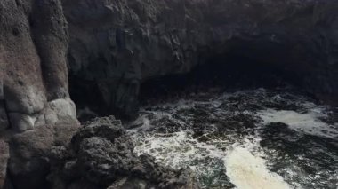 Ağır çekim bir video, karanlık volkanik kayalar tarafından çerçevelenmiş bir mağaraya yavaşça giren okyanus dalgalarını yakalar..