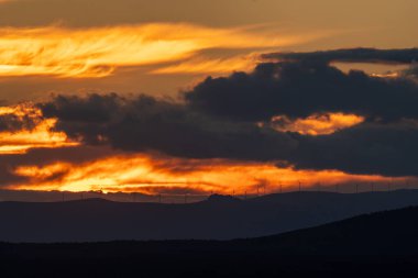 A fiery sunset sky highlights wind turbine silhouettes on a distant hill, creating a striking and peaceful landscape.