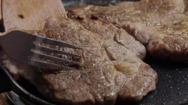 Bir çatal ve ahşap bir spatulayla ustalıkla pişirilmiş iki sulu, mükemmel kurutulmuş bifteğin yakın çekim görüntüsü.