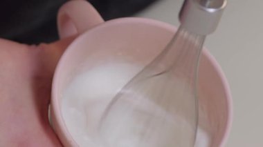 Bir el pembe bardakta krema çırpıyor, dokusunu ve bütün karıştırma sürecini güzelce gösteriyor.