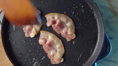 Domuz pastırması sıcak tavada cızırdıyor. Mükemmel pişirme sonuçları elde etmek için ahşap kaşıkla ustalıkla ters çevrilmiş.