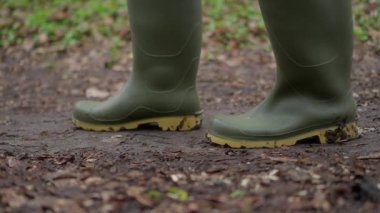 Yeşil Wellington çizmeleri giyen bir insan, verimli ve canlı doğayla çevrili çamurlu bir patikada rahatça yürür.