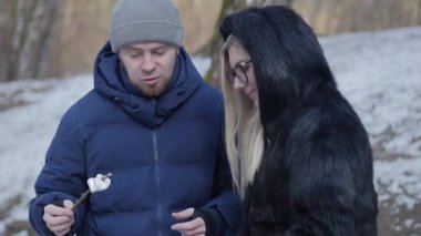 Bir çift, güzel, karlı bir manzaraya yuva yapmış lezzetli marşmelovları neşeyle kızartıyorlar.