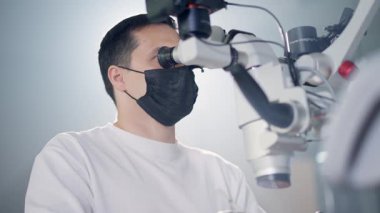 Modern stomatoloji kliniğinde diş mikroskobuyla çalışan erkek dişçi. Doktor kanal tedavisi için mikroskop kullanıyor. Stomatolog cerrahi bir operasyon gerçekleştirir. Kozmetik dişçiliği ve ameliyat. Yüksek kalite 4k görüntü
