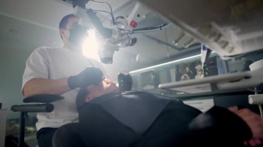 Modern stomatoloji kliniğinde diş mikroskobuyla çalışan erkek dişçi. Doktor kanal tedavisi için mikroskop kullanıyor. Stomatolog cerrahi bir operasyon gerçekleştirir. Kozmetik dişçiliği ve ameliyat. Yüksek kalite 4k görüntü