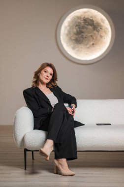 Bir kadın, parlak bir dolunayın önünde beyaz bir kanepede rahat bir şekilde oturuyor ve rahat oturma odasında güzel bir sahne yaratıyor.