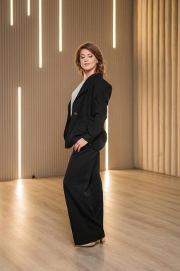 Şık siyah takım elbiseli profesyonel bir kadın, kendine güvenen bir şekilde dekore edilmiş bir odada, resmi kıyafetlerini sergiliyor.