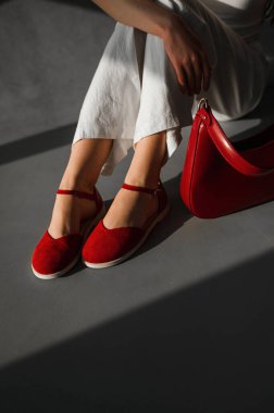 Bir kadın şık kırmızı ayakkabılar giyip yanında kırmızı bir çanta taşırken yerde rahatça oturuyor.