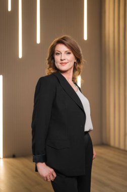 Zarif, siyah bir takım elbise giymiş bir kadın, kendine güvenen bir şekilde iyi aydınlatılmış bir odada duruyor, profesyonellik ve özgüven saçıyor.