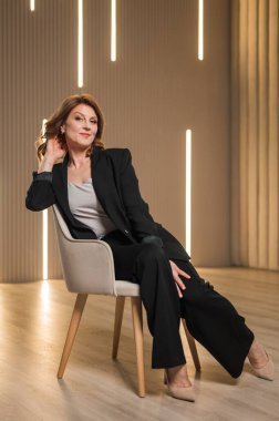 Şık giyinmiş profesyonel bir kadın koltukta rahat bir şekilde oturur bacaklarını bağlar, kendine güveni ve duruşuyla.