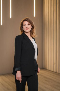 Zarif, siyah bir takım elbise giymiş bir kadın, kendine güvenen bir şekilde iyi aydınlatılmış bir odada duruyor, profesyonellik ve özgüven saçıyor.