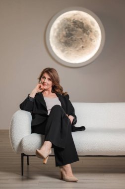 Pürüzsüz siyah takım elbiseli bir kadın şık beyaz bir koltukta rahatça oturuyor ve rahat ama profesyonel bir imajı canlandırıyor.