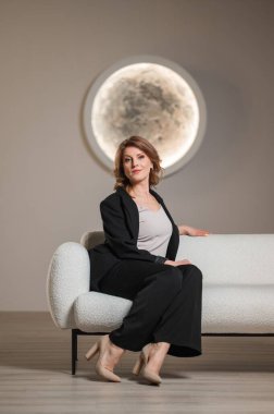 Pürüzsüz siyah takım elbiseli bir kadın şık beyaz bir koltukta rahatça oturuyor ve rahat ama profesyonel bir imajı canlandırıyor.