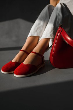 Bir kadın şık kırmızı ayakkabılar giyip yanında kırmızı bir çanta taşırken yerde rahatça oturuyor.