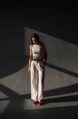 Bir kadın zarif bir şekilde karanlık bir odada dikiliyor, beyaz bir bluz giyiyor, aynı beyaz pantolona sahip, çarpıcı bir kontrast oluşturuyor.
