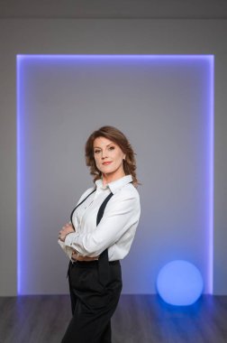 Gıcır gıcır beyaz gömlekli ve şık siyah pantolonlu bir kadın, kollarını kavuşturmuş özgüvenle iyi aydınlatılmış bir odada dikiliyor.