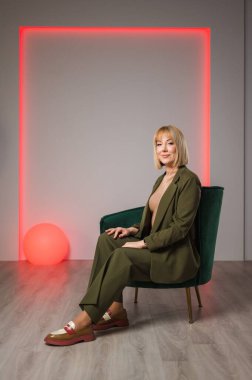 Şık bir takım elbise giymiş bir kadın zarif yeşil bir sandalyeye oturtulur, kolları düşünceli bir şekilde kavuşturulur.