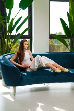 Bir kadın rahat bir şekilde büyük bir pencerenin önündeki rahat, mavi bir koltukta oturuyor, odaları doğal ışık arttırıyor.