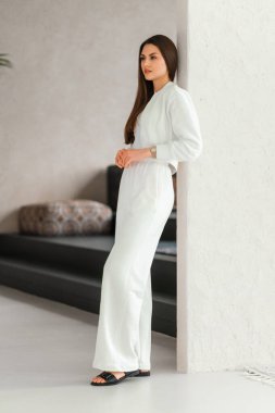 Şık, beyaz bir elbise giymiş bir kadın nazikçe duvara yaslanıyor ve zarafetin çarpıcı bir görsel izlenimini yaratıyor.