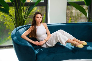 Bir kadın rahat bir şekilde büyük bir pencerenin önündeki rahat, mavi bir koltukta oturuyor, odaları doğal ışık arttırıyor.