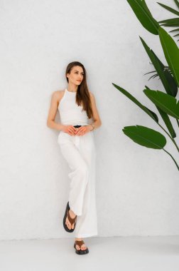 Şık beyaz tulum giymiş bir kadın sahneyi güzelleştiren güzel bir saksı bitkisinin yanında zarif bir şekilde duruyor.