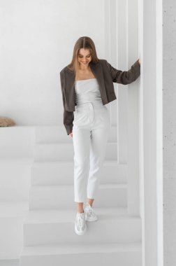 Moda düşkünü genç bir kadın modern, minimalist bir alanda poz veriyor. Kırpılmış ceketi ve beyaz pantolonu olan şık bir kıyafeti sergiliyor. Yaşam tarzı ve moda temaları için mükemmel.