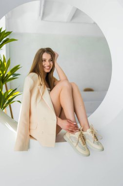 Şık bir genç kadın zarif bir şekilde krem rengi ceket ve spor ayakkabılarla poz veriyor, çağdaş bir iç dekorda dengeli bir zarafet ve moda karışımı sergiliyor..