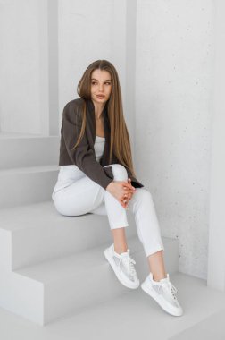 Uzun saçlı genç bir kadın beyaz merdivenlerde zarif bir şekilde oturuyor. Üstü açık bluz, ceket ve beyaz spor ayakkabılar giyiyor. Modern, minimalist bir estetik....