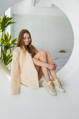 Şık bir genç kadın zarif bir şekilde krem rengi ceket ve spor ayakkabılarla poz veriyor, çağdaş bir iç dekorda dengeli bir zarafet ve moda karışımı sergiliyor..