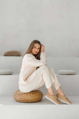 Tarafsız bir kıyafet giymiş zarif bir kadın örülmüş bir tabureye oturuyor. Huzurlu, minimalist arka plan rahatlatıcı havasını arttırır..