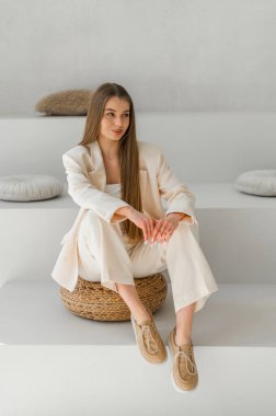 Tarafsız bir kıyafet giymiş zarif bir kadın örülmüş bir tabureye oturuyor. Huzurlu, minimalist arka plan rahatlatıcı havasını arttırır..