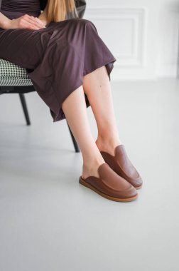 Bir çift şık kahverengi terlik modern bir iç mekanda sergilenen şık bir giysinin yanında sergileniyor, moda ve yaşam tarzı tasvirleri için mükemmel..