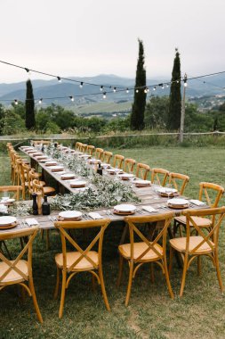 A beautifully decorated rustic wedding table setup with greenery garlands, white plates, and elegant glassware in the serene Tuscany countryside. clipart