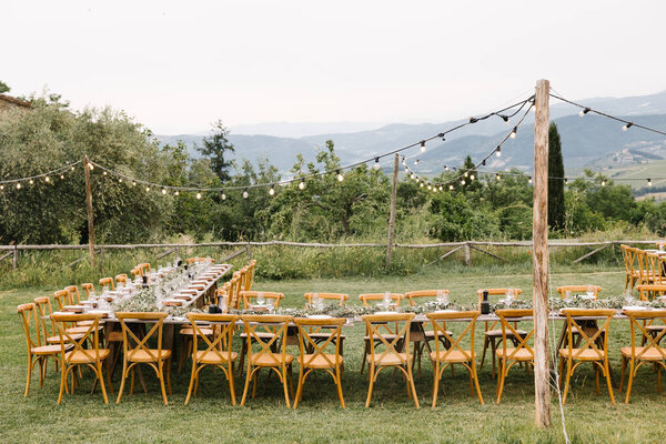 Элегантный открытый свадебный прием в спокойной тосканской сельской местности, с длинными деревянными столами, украшенными зеленью и струнными огнями.