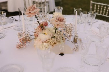 Göz kamaştırıcı bir düğün masası. Resimli bir kırsal alana kurulmuş. Düzenlemede pampalar çimenler, beyaz ve allık güller yer alıyor.