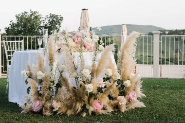 Göz kamaştırıcı bir düğün masası. Resimli bir kırsal alana kurulmuş. Düzenlemede pampalar çimenler, beyaz ve allık güller yer alıyor.