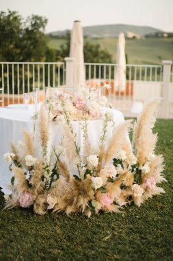 Göz kamaştırıcı bir düğün masası. Resimli bir kırsal alana kurulmuş. Düzenlemede pampalar çimenler, beyaz ve allık güller yer alıyor.