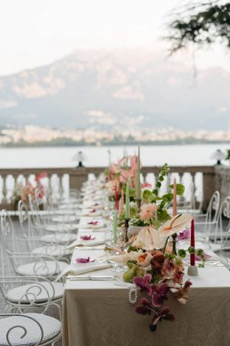 Como Gölü 'ne bakan terasta güzel dekore edilmiş bir açık hava yemek masası. Tasarımda, renkli mumlar ve beyaz işlenmiş demir çayla vurgulanan yumuşak pastel tonlarda orkideler, anthuriumlar ve yıldız çiçekleriyle hassas çiçek düzenlemeleri yer alıyor.