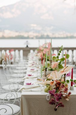 Como Gölü 'ne bakan terasta güzel dekore edilmiş bir açık hava yemek masası. Tasarımda, renkli mumlar ve beyaz işlenmiş demir çayla vurgulanan yumuşak pastel tonlarda orkideler, anthuriumlar ve yıldız çiçekleriyle hassas çiçek düzenlemeleri yer alıyor.