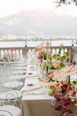 Como Gölü 'ne bakan terasta güzel dekore edilmiş bir açık hava yemek masası. Tasarımda, renkli mumlar ve beyaz işlenmiş demir çayla vurgulanan yumuşak pastel tonlarda orkideler, anthuriumlar ve yıldız çiçekleriyle hassas çiçek düzenlemeleri yer alıyor.