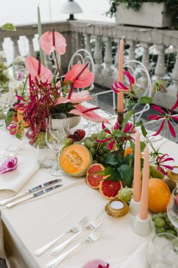 A beautifully decorated outdoor dinner table set on a terrace overlooking Lake Como. The design features delicate floral arrangements with orchids, anthuriums, and dahlias in soft pastel tones, accented by colorful candles and white wrought iron chai clipart