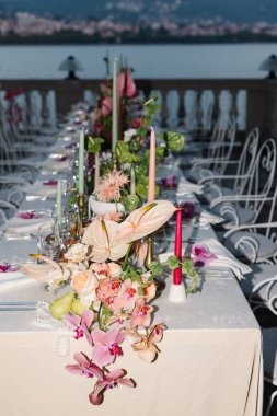 A luxurious wedding table setup overlooking the serene waters of Lake Como. The design features vibrant pink anthuriums, tropical fruits such as grapefruits, kiwis, papayas, and pomegranates, accompanied by greenery and fuchsia gloriosa flowers. clipart