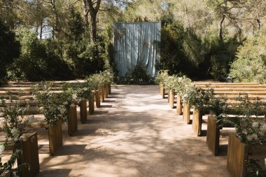 Sakin bir orman ortamında güzel bir açık hava düğün töreni düzenlendi. Ahşap banklar koridor boyunca uzanır, yemyeşil çiçeklerle süslenir ve zarif çiçeklerle süslenir.