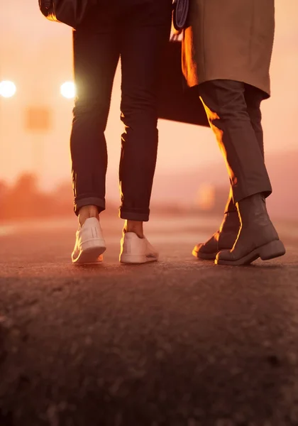 Legs of a man and woman with bags walk embraced on a wet road with a street lamp at sunset. Rear view. 3D render.